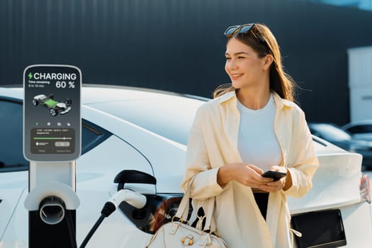 Young woman holding shopping bag and use smartphone to pay for electricity for recharging EV car battery from charging station at city mall parking lot. Modern woman go shopping by eco car. Expedient