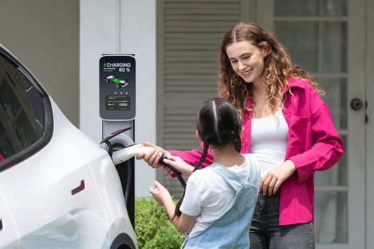 Happy little young girl learn about eco-friendly and energy sustainability as she help her mother recharge electric vehicle from home EV charging station. EV car and modern family concept. Synchronos