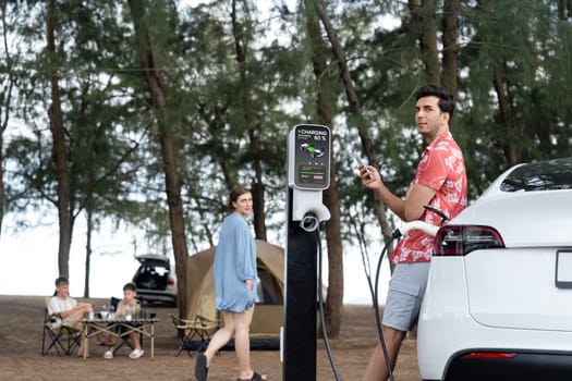 Outdoor adventure and family vacation camping in nature travel by eco friendly car for sustainable future. Lovely sit on trunk, charging EV car with EV charging station in campsite. Perpetual