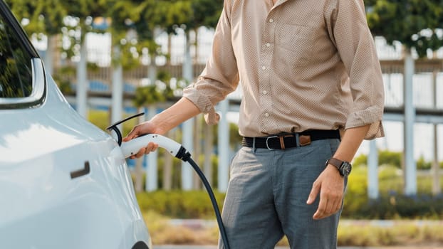 Young man put EV charger to recharge electric car's battery from charging station in city commercial parking lot. Rechargeable EV car for sustainable environmental friendly urban travel. Expedient