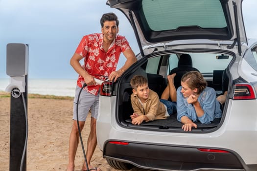 Family vacation trip traveling by the beach with electric car, lovely family sit on the trunk, charging EV car battery with green and sustainable energy. Family travel and eco-friendly car. Perpetual