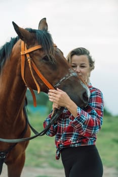 Happy blonde with horse in forest. Woman and a horse walking through the field during the day. Dressed in a plaid shirt and black leggings