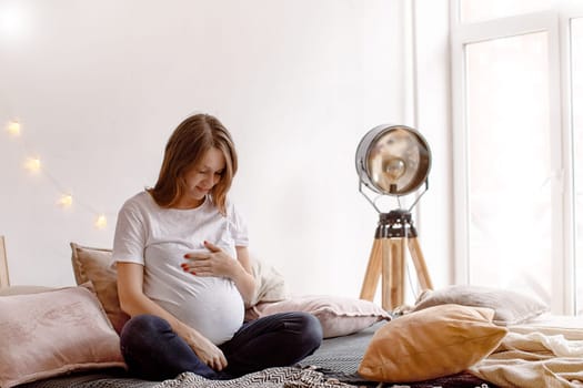 Beautiful pregnant lady smiling and touching belly while sitting on comfortable bed in cozy room