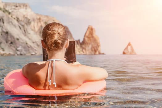 Woman freelancer works on laptop swimming in sea on pink inflatable ring. Pretty lady typing on computer while floating in the sea on inflatable donut at sunset. Freelance, remote work on vacation