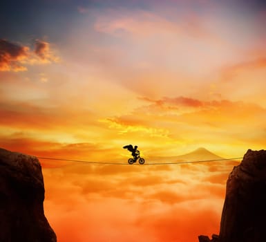 Boy with angel wings balance on a rope over a chasm riding a bicycle.Self overcoming and risk taking concept. Beautiful sunset background over the clouds