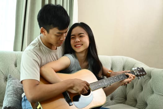 Shot of loving young couple sitting on couch and man teaching his girlfriend playing guitar.