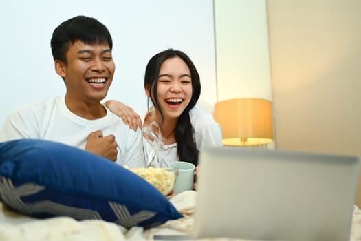 Happy young couple watching movie on laptop and eating popcorn in their bedroom.