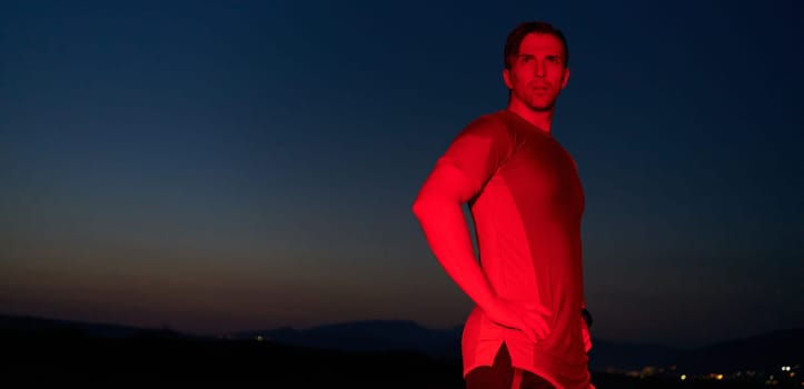 In the solemn darkness illuminated by a red glow, an athlete strikes a confident pose, embodying resilience and determination after completing a grueling day-long marathon.