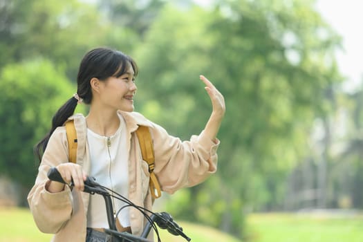 Cheerful young female student with backpack riding bicycle and waving hand as greeting someone.