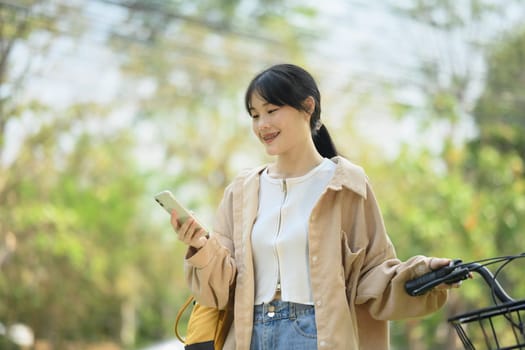 Positive young woman standing near bicycle and using mobile phone, communication online.