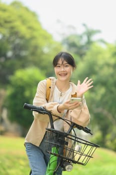 Portrait of cheerful young female student sitting on bicycle and using mobile phone.