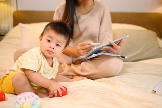 Little baby boy playing with toys and mother working with digital tablet on bed. Motherhood and child care concept.