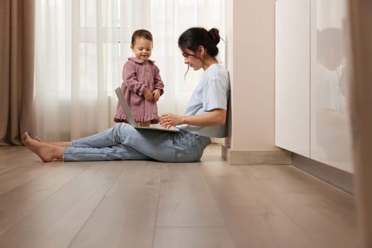 beautiful casual woman sitting on the floor and working on laptop with her little child girl at home