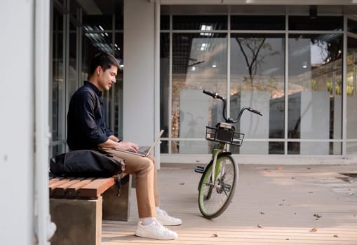 A young Asian businessman rides a bicycle to work. Use a reusable water bottle. Working outside of the office using a laptop The concept of saving energy and reducing pollution to the environment..