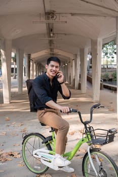 A young Asian businessman rides a bicycle to work. Standing outside the office talking on the phone and talking about business. Concept of reducing energy and reducing air pollution..