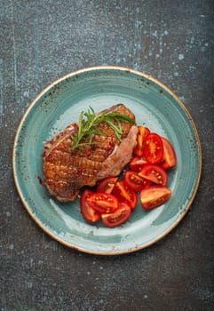 Delicious roasted duck breast fillet with golden crispy skin, with pepper and rosemary, top view on ceramic blue plate served with cherry tomatoes salad, rustic concrete rustic background