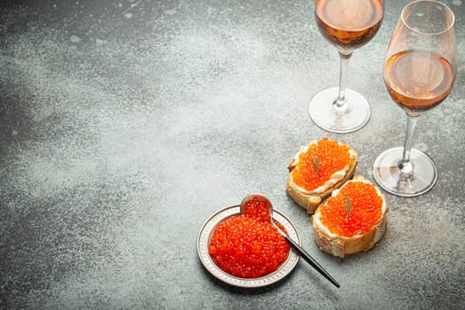 Small metal plate with red salmon caviar, two caviar toasts canape, two glasses of champagne top view on grey concrete background, festive luxury delicacy and appetizer. Space for text.