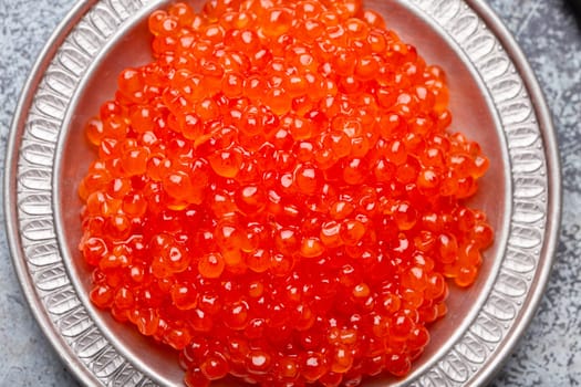 Small metal plate with red salmon caviar and a spoon top view on grey concrete background, festive luxury delicacy and appetizer.