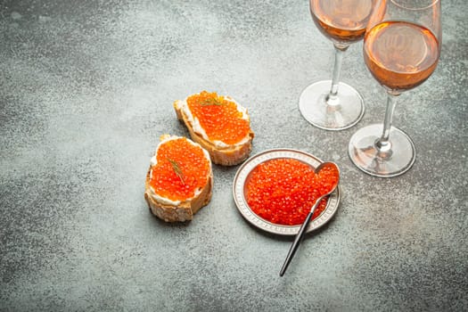 Small metal plate with red salmon caviar, two caviar toasts canape, two glasses of champagne top view on grey concrete background, festive luxury delicacy and appetizer.