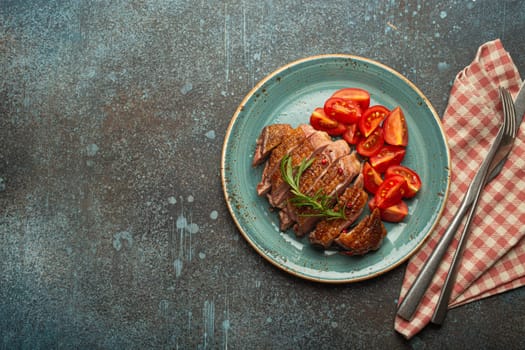 Delicious roasted sliced duck breast fillet with golden crispy skin, with pepper and rosemary, top view ceramic blue plate served with cherry tomatoes salad, concrete rustic background, copy space.