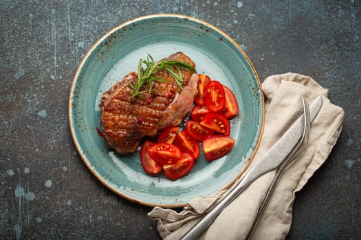 Delicious roasted duck breast fillet with golden crispy skin, with pepper and rosemary, top view on ceramic blue plate served with cherry tomatoes salad, rustic concrete rustic background