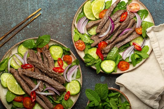 Two plates with traditional Thai beef salad with vegetables and mint top view served on rustic concrete background, healthy exotic asian meal.