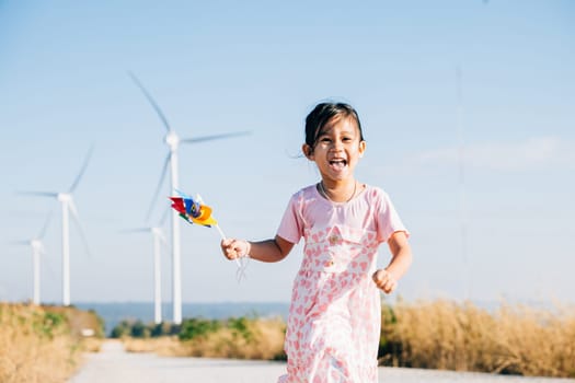 A joyful child holding pinwheels runs near windmills embodying wind energy education. Clean electricity and sustainable industry visuals in a picturesque wind turbine landscape.