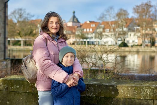 Riverside Family Harmony: Mother, 30 Years Old, and Son - Beautiful 8-Year-Old Boy, Standing by Neckar River and Historic Half-Timbered Town, Bietigheim-Bissingen, Germany, Autumn. Immerse yourself in the picturesque harmony of familial bonds by the riverside with this captivating image. A mother, 30 years old, and her son - a beautiful 8-year-old boy, standing by Neckar River and the historic half-timbered town of Bietigheim-Bissingen, Germany. The autumnal colors add warmth to this timeless family moment.