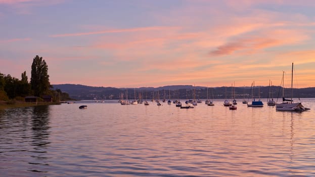 Bodensee Lake Sunrise Panorama. Morning Sunlight Over Tranquil Waters. Witness the mesmerizing dawn over Germany's Bodensee Lake, captured from a boat dock. Embrace the tranquil beauty of the early morning as the sun rises, casting a soft glow on the landscape. The peaceful scene features boats, yachts, and a charming water shack set against a backdrop of a captivating sky. Clouds delicately reflect on the calm water, creating a serene atmosphere. Immerse yourself in the serene beauty of a lakeside sunrise. Explore the harmony of nature, technology, and production as the day unfolds by the lake.