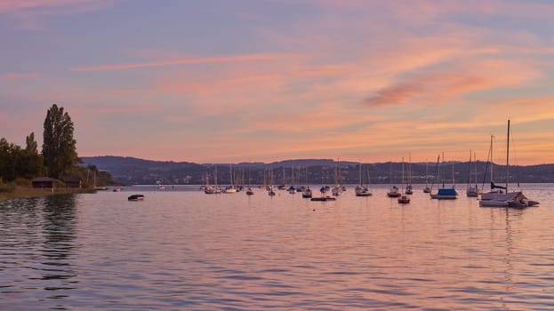 Bodensee Lake Panorama. Evening, twilight, setting sun, picturesque landscape, serene waters, boats and yachts at the dock, beautiful sky with clouds reflecting in the water, riverside at dusk, showcasing the coexistence of technology and production with the environment.