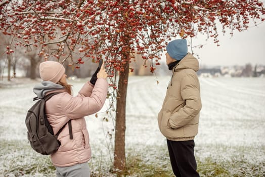 Winter Romance: Girl in Pink Winter Jacket Photographing Boy Against Snow-Covered Red Tree and Field. Embrace the winter magic in this enchanting image, where a girl in a pink winter jacket captures a moment as she photographs her companion against the backdrop of a snow-covered red tree and field. The photograph beautifully conveys the essence of winter romance and the serene beauty of a snowy landscape.