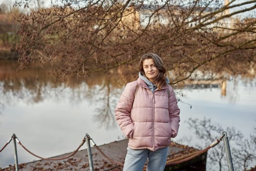 A young woman standing at the shore looking at the river in autumn sunny day. Street view, copy space for text, travel photo. Happy tourist woman on the bank of the river in autumn in warm clothes. Tourists enjoy their vacation, winter season. Romantic look and travel concept. A joyful mood in a Caucasian girl. Winter Wonderland: Enchanting Girl by the Riverside in Autumn.