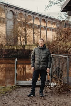 Timeless Elegance: 40-Year-Old Man in Stylish Jacket by Neckar River and Historic Bridge in Bietigheim-Bissingen, Germany. Experience the allure of seasons as a charismatic 40-year-old man stands gracefully by the enchanting Neckar River, adorned in a sophisticated jacket, against the backdrop of historic bridge pillars in the charming city of Bietigheim-Bissingen, Germany. This captivating image seamlessly blends timeless elegance with the scenic beauty of autumn or winter, creating a picturesque scene of urban exploration and mature style in the heart of German heritage