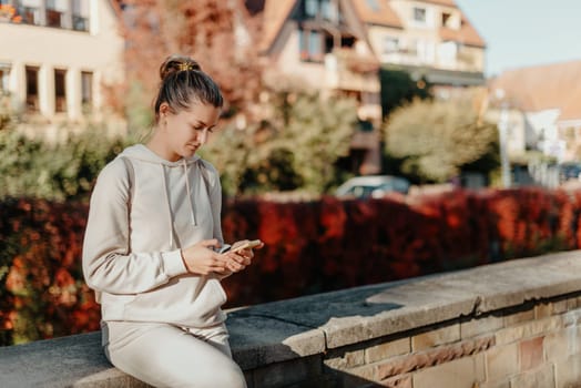 Young fashionable teenage girl with smartphone in park in autumn sitting at smiling. Trendy young woman in fall in park texting. Retouched, vibrant colors. Beautiful blonde teenage girl wearing casual modern autumn outfit sitting in park in autumn. Retouched, vibrant colors, brownish tones.