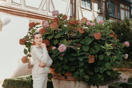 Attractive curly blonde woman walk on the city park street. Girl wear purple hoodie look happy and smiles. Woman make here me gesture standing near pink blooming bush flowers. Happy laughing girl