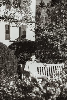 a teenager sits on a bench in the park drinks coffee from a thermo mug and looks into a phone. Portrait of handsome cheerful guy sitting on bench fresh air using device browsing media smm drinking latte urban outside outdoor.