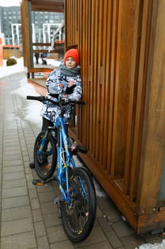 A Caucasian boy dressed in a winter jacket walks with a bicycle around the city.