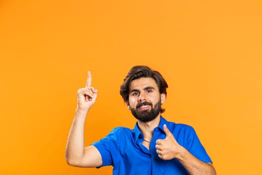 Young man showing thumbs up and pointing empty place above head, advertising area for commercial text, copy space for goods promotion advertisement. Guy isolated on orange studio background. Lifestyle