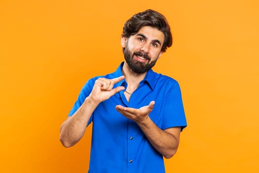 Need some more, please give me, help. Man showing a little bit gesture with sceptic smile, showing minimum sign measuring small size. Middle eastern guy isolated alone on orange studio background