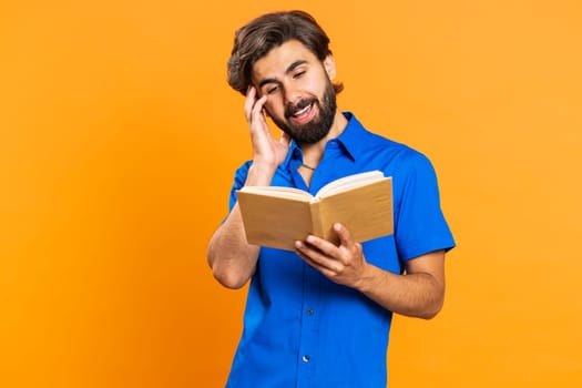 Young excited middle eastern man reading funny interesting fairytale story book, laughing, leisure hobby, knowledge wisdom, education, learning study, wow. Guy isolated on orange studio background