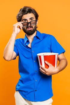 Excited young man in 3D glasses eating popcorn and watching interesting tv serial, sport game, film, online social media movie content. Middle eastern guy in shirt isolated on orange studio background