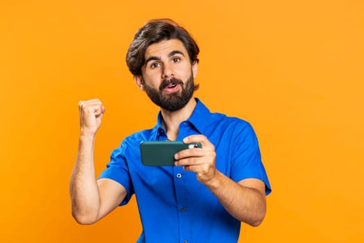Worried funny addicted man enthusiastically playing racing video games on mobile phone. Happy middle eastern guy using smartphone gadget app with drive simulator isolated on orange studio background