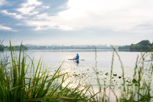 A man kayaks among water lilies on a calm lake, against the backdrop of a cityscape shrouded in fog