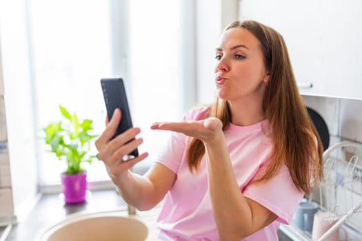 Woman blowing kiss at smartphone for video call. Modern communication and technology concept. Casual home lifestyle with copy space. Design for social media, banner.