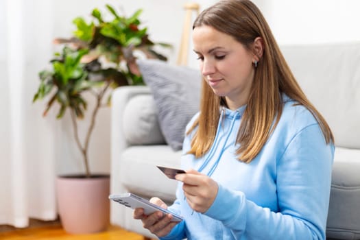 Focused woman using smartphone and credit card for online shopping, sitting on a couch at home.