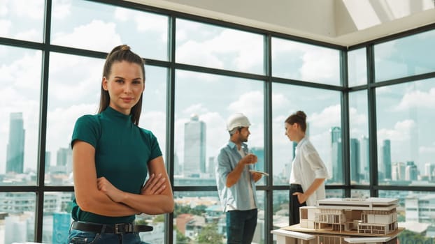 Young beautiful interior designer crossing arms while engineer team talking about house design. Skilled businesswoman smiling at camera with confident while sitting near house model. Tracery