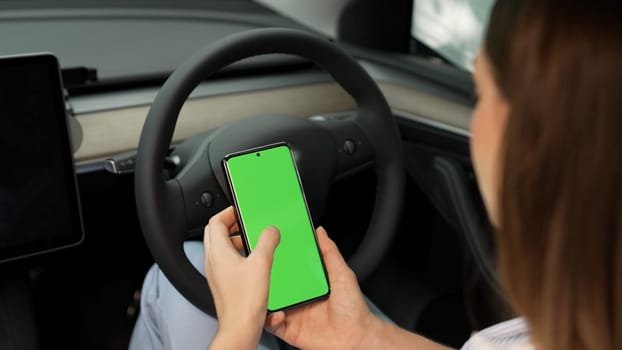 Holiday vacation road trip with environmental-friendly car concept. Eco-conscious woman on driver seat holding blank copyspace green screen smartphone for EV battery status. Exalt