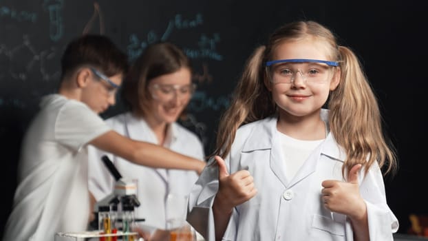 Smart caucasian girl showing thumb while teacher and student doing experiment behind. Child looking at camera while boy looking under microscope at black board written chemical theory. Erudition.