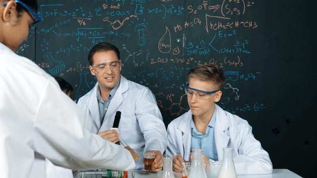 Creative teacher pointing chemistry at blackboard while talking to highschool boy at table with microscope and test tube with colored liquid. Young student wearing lab coat in STEM class. Edification.