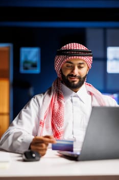 A focused Arabian businessman in Islamic attire sits at a modern desk, typing on a laptop. He efficiently works, utilizing wireless technology for online communication, surfing the net, and conducting online shopping and payments.